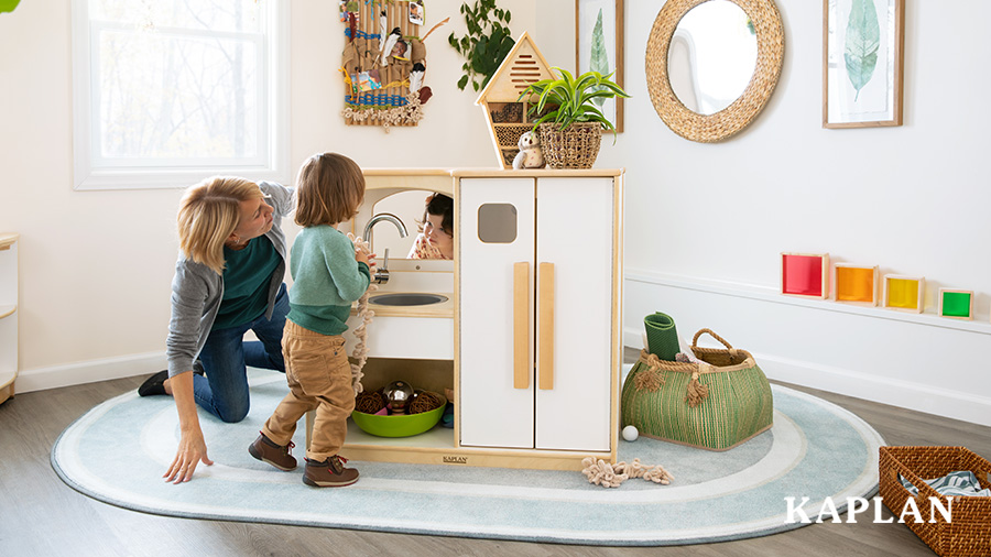 A teacher and two toddler students play in a white and natural wooden kitchen set that is sitting a light blue, oval shaped rug, in a natured based classroom.