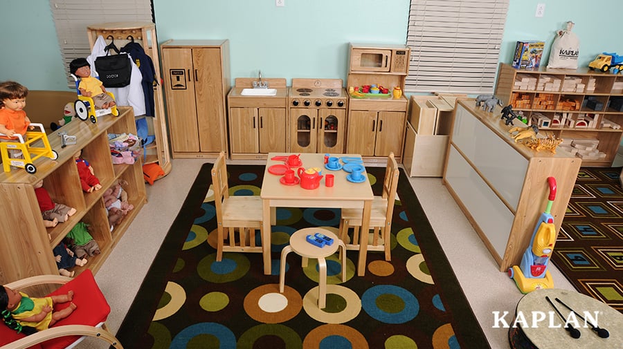 Wooden kitchen furniture in a dramatic play center filled with toys, sits on a large, rectangular, tan, brown, green and blue, geometric rug.