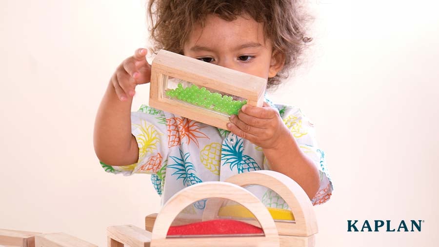 A two-year-old child in a Hawaiian shirt with wavy hair plays with Kaplan Rainbow Sensory Blocks.
