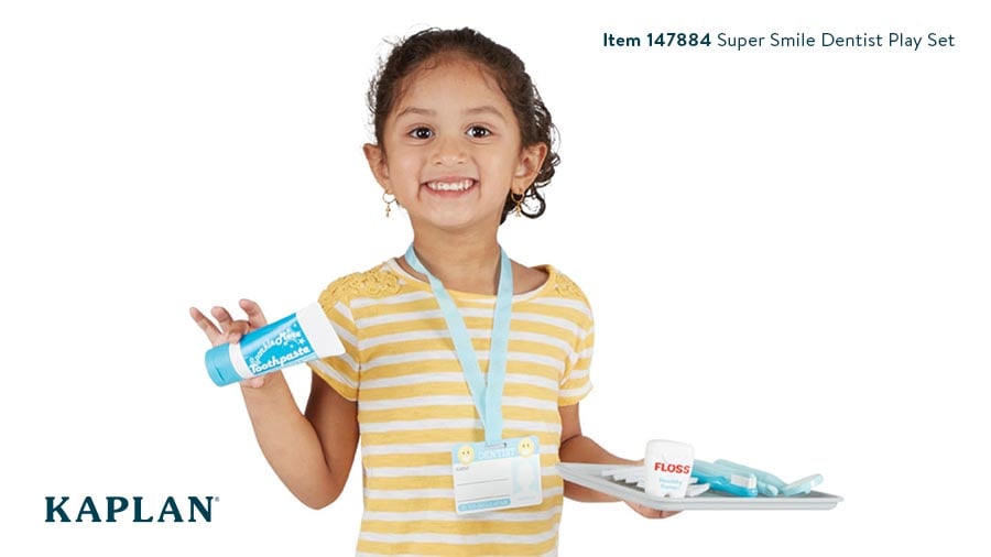 A young girl in a yellow t-shirt holds a tray of dental dramatic play toys and a tube of pretend toothpaste.