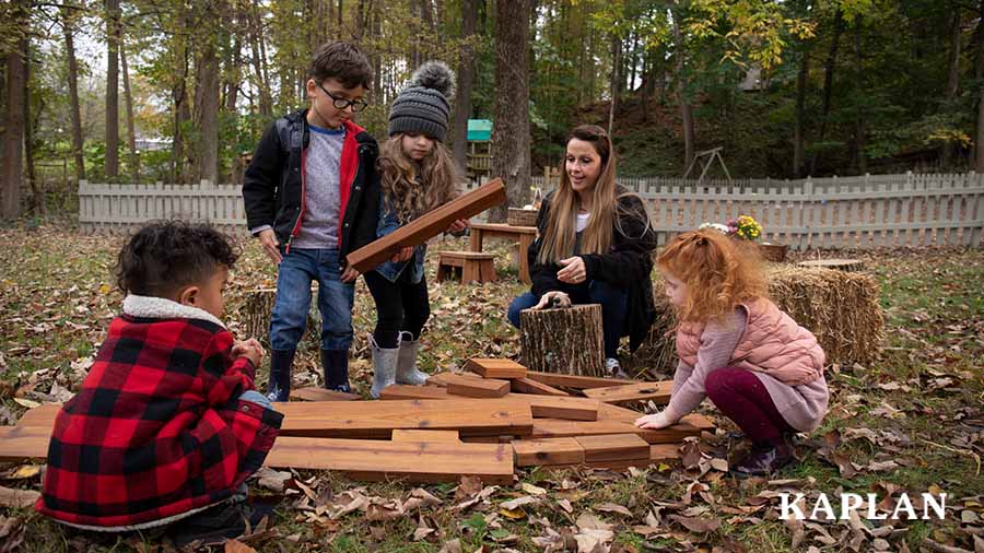 Four young children gather around a pile of thermally modified wooden boards on their outdoor playground, while a female teacher sits nearby, watching them play and build with the wooden boards. 