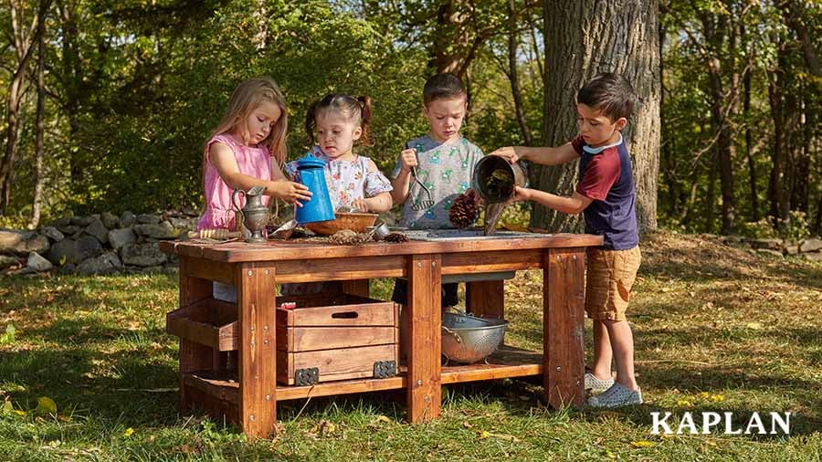 Four young children are using pots, pans, and pinecones to create a messy meal on the Nature to Play Mud Kitchen located in an outdoor play space.