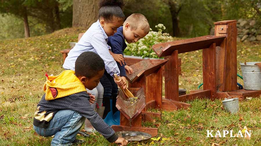 Three children in an outdoor play space use their hands and wooden spoons to manipulate the flow of water on the Nature to Play Water Trough System.