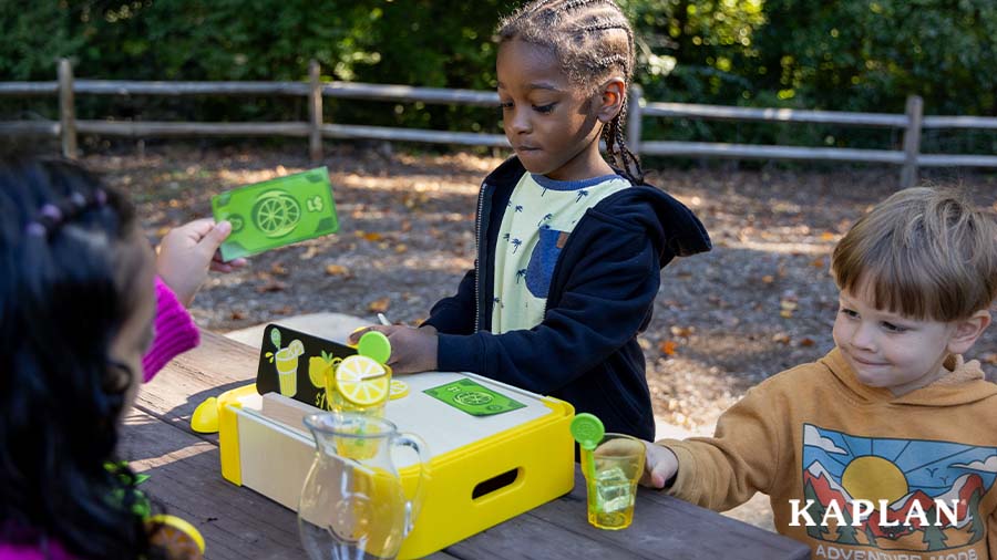 Three young children sit outside at a picnic table, the Lemonade Stand Set is in front of them. One child is slicing a pretend lemon, another is holding a plastic glass of pretend lemonade, and the third is holding a pretend $1 bill up in the air. 