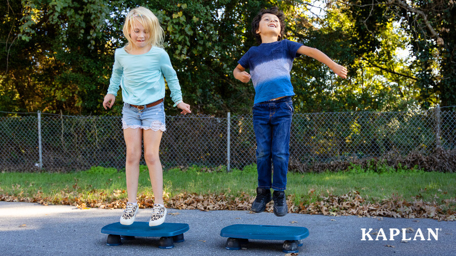 Two children smile as they jump on two blue Bouncy Boards in an outdoor area. 