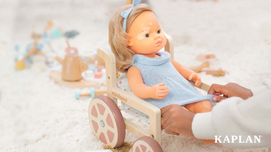 An image of a young child's hands pulling down a dress to cover the Miniland Doll with Vitiligo's legs. The doll is sitting in a wooden Miniland Doll Wheelchair. 
