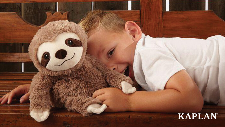 A young child lays on a wooden bench with a Warmies Microwavable Plush Sloth toy. 