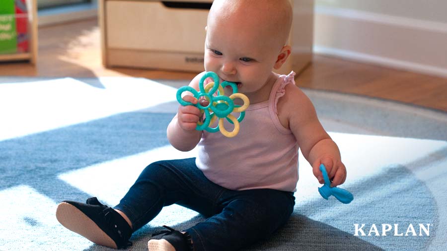 A young child holds a green, yellow, and blue Silicone Fun Loop in both hands. She is lifting the rings in her right hand up to her mouth as if to chew on the rings. 