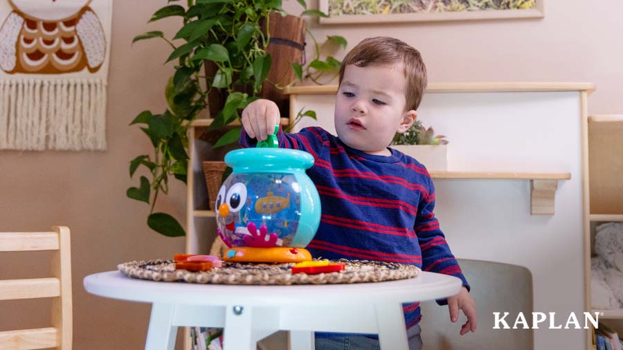A young child sits in a playroom, a Color Fun Fishbowl is in front of the child on a round wooden table. The child is placing a green fish piece inside the fishbowl. 