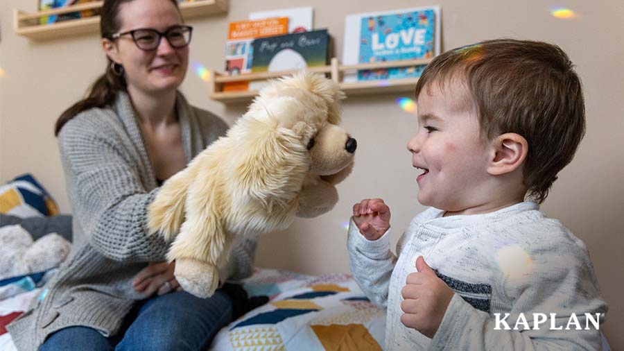 A woman is holding a Golden Retriever Puppet in front of a young boy. The boy is looking at the puppet and smiling. 