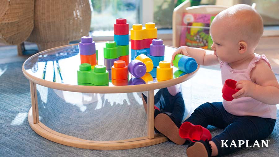 A young child sits beside a Reflection Table, a set of Soft Stack Baby Bricks are on top of the table. The child is reaching out for a green and blue block.