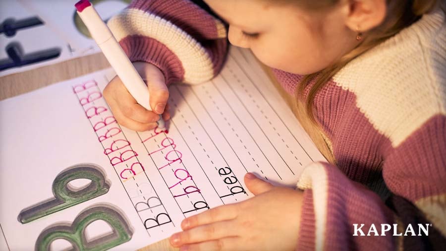 A young child practices writing the letter B on a lined worksheet using a pink marker