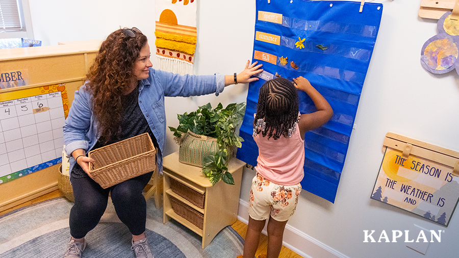 An early childhood teacher sits in a wooden chair for a circle time activity, a young girl is standing beside the teacher and is placing a leaf in a pocket chart which is labeled with seasonal words. 