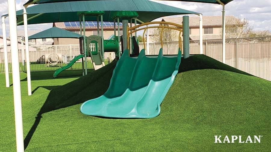 A green slide with three sections underneath a shade on a playground with green synthetic turf surfacing