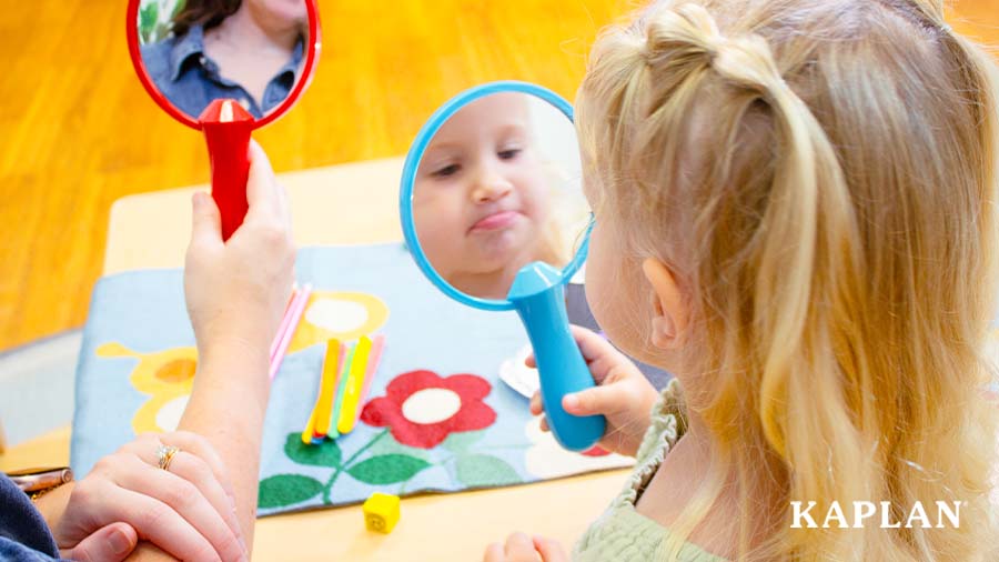 A young child is holding a blue hand mirror, she is looking at her reflection and making a sad face. 