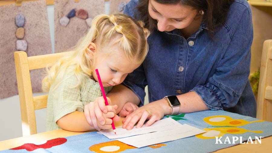 An early childhood teacher sits beside a young child, the teacher is helping the child write on a sheet of paper which is laying on the table in front of the child. 