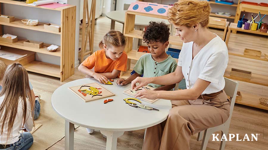 An early childhood teacher sits at a table with two children, the three of them are adding pieces to complete a wooden fish puzzle. 