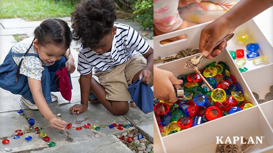 Two young children use tweezers to collect colorful gems and brass keys on a stone landing