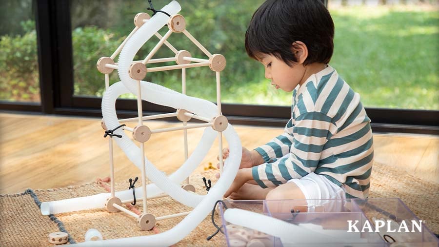A preschooler builds a tall structure using wooden rods and connectors, with flexible white tubes weaved throughout