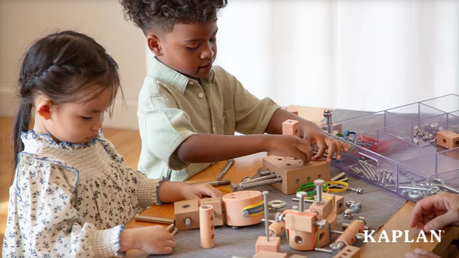 Two preschoolers build with wooden blocks, metal bolts, and rubber bands at a table