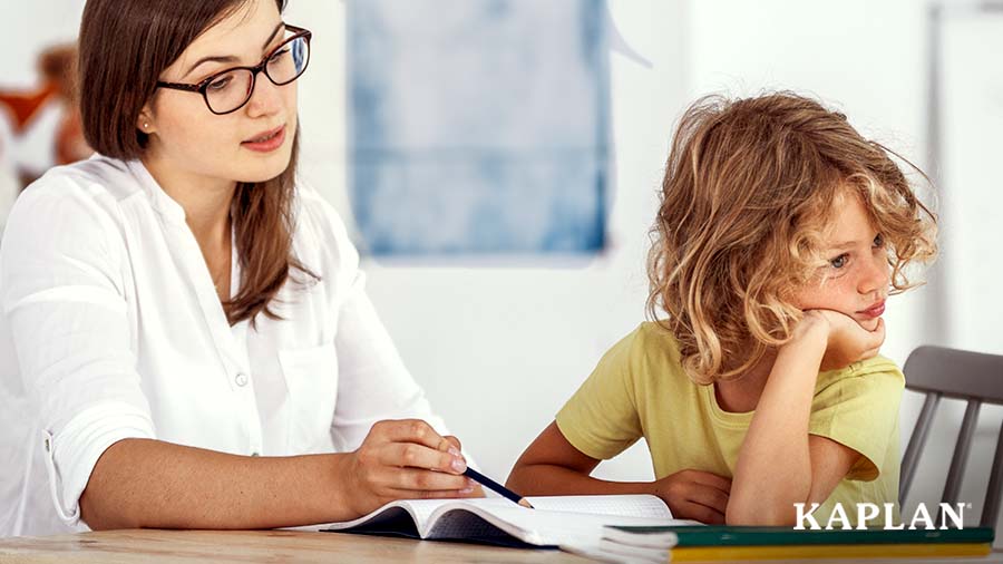 A woman in a white shirt sits at a table with a child in a yellow shirt. The child is looking away from the woman while the woman is pointing to something in a book. 
