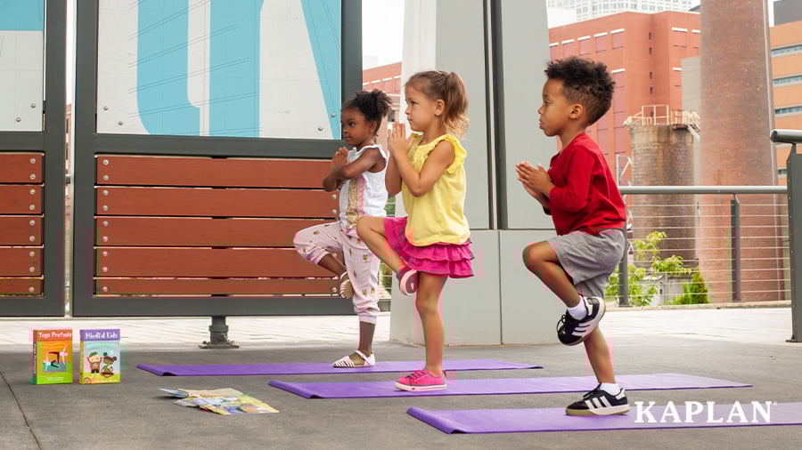 Three children stand on purple yoga mats while doing a tree pose. 
