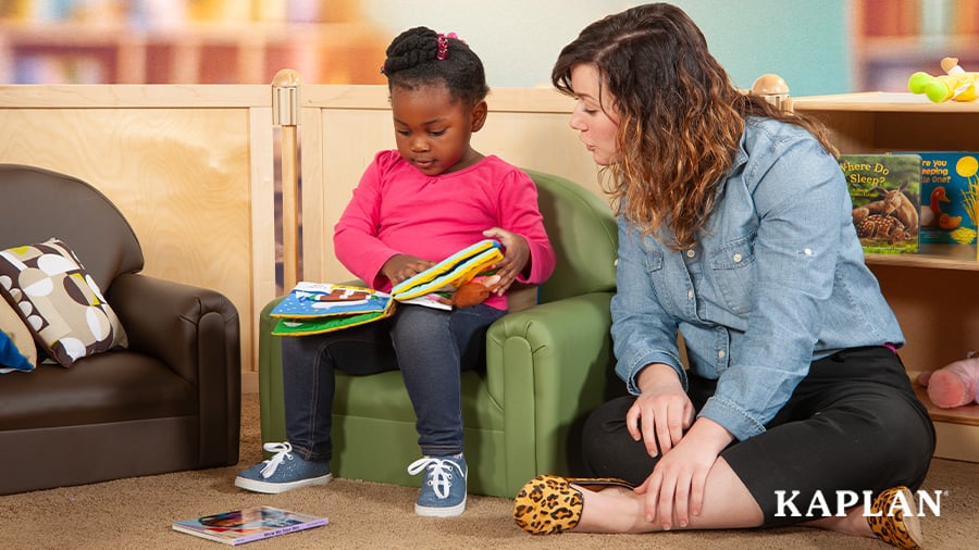 An early childhood teacher sits beside a child who is sitting in a child-sized green chair reading a soft covered book. 
