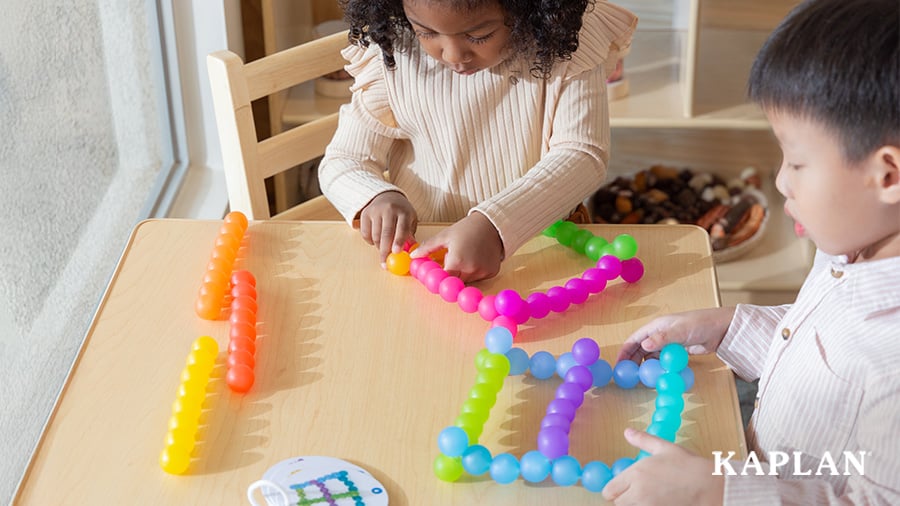 Two children sit at a wooden table while playing with a set of Kaplan's Bendi Beads. 