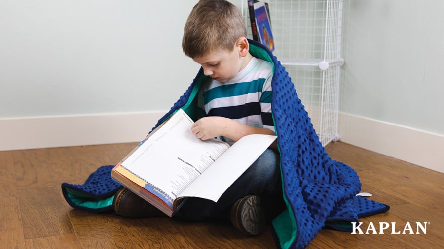 A child sits on the floor, covered with a weighted blanket as they flip through the pages of a large book. 