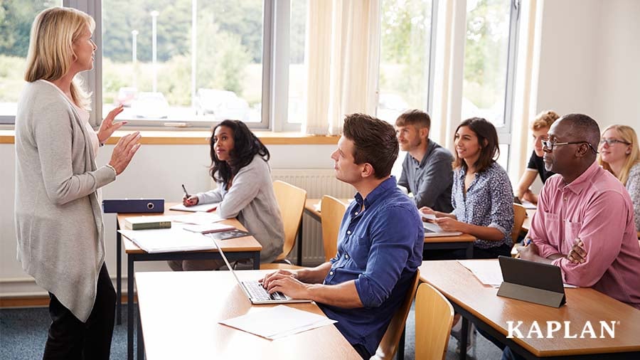 A group of early childhood educators sit at desks in a room, looking up toward the front of the room at an expert trainer sharing knowledge on early childhood topics. 