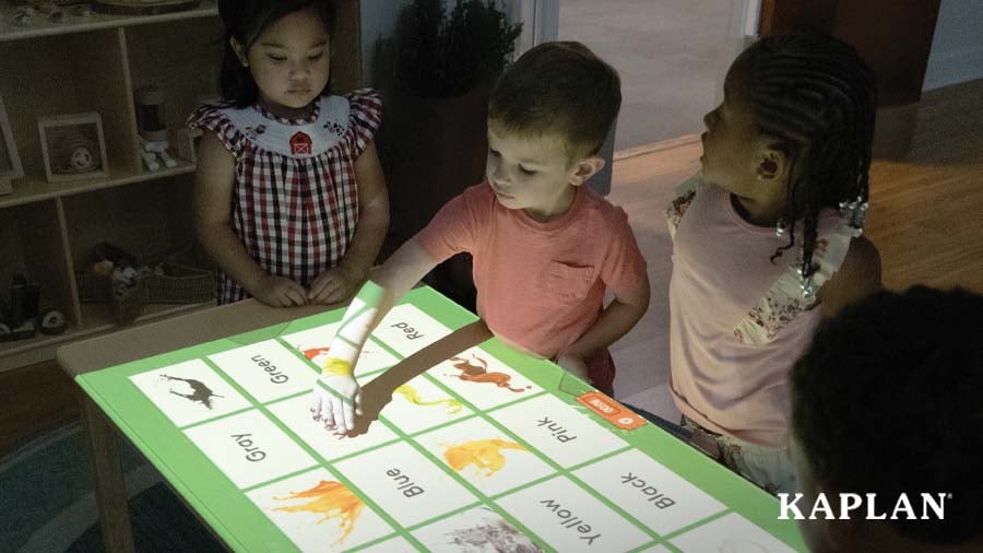 A preschool-aged boy interacts with a digital display projected on a table while two preschool-aged girls stand beside him and watch