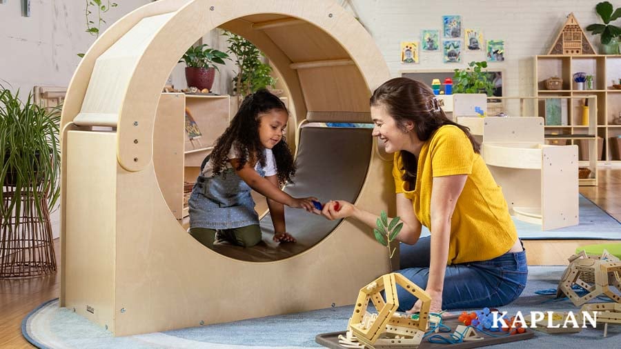 An early childhood educator sits on the floor and smiles while handing a loose parts toy to a young child who is sitting inside the Carolina Connections Cove. 