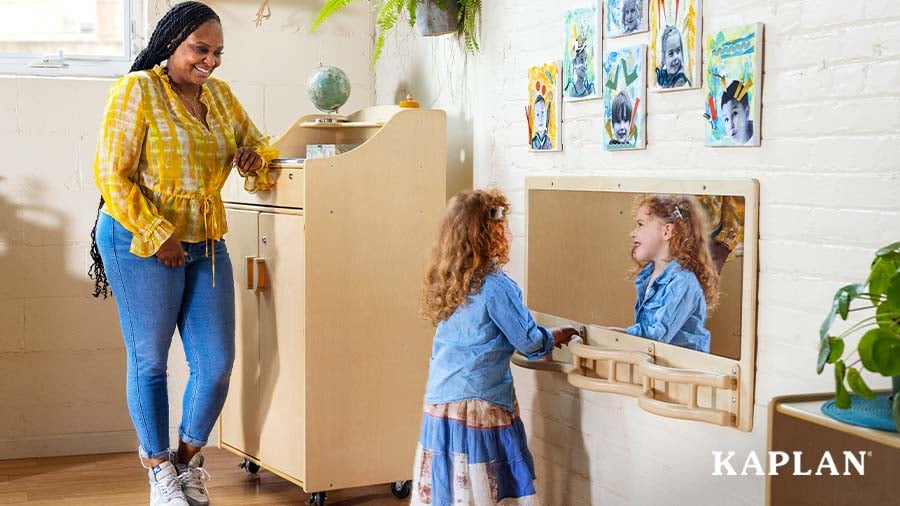 A teacher smiles and watches a young child ambulate through the classroom by holding onto the Carolina Connections Explorer Rail with attached Mirror.