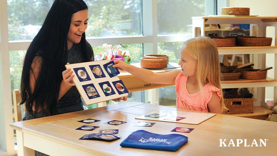 An early childhood teacher sits with a young child as she completes a puzzle activity. The teacher is smiling at the child. 