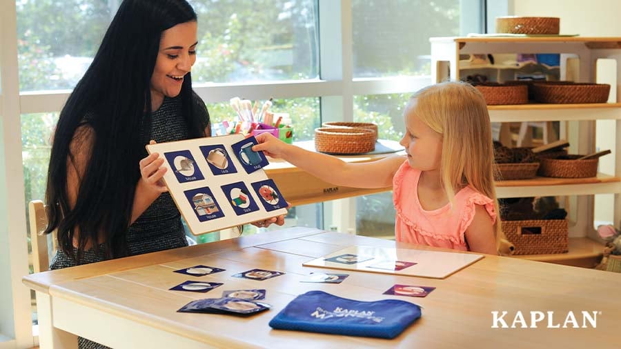 An early childhood teacher sits with a young child as she completes a puzzle activity. The teacher is smiling at the child.