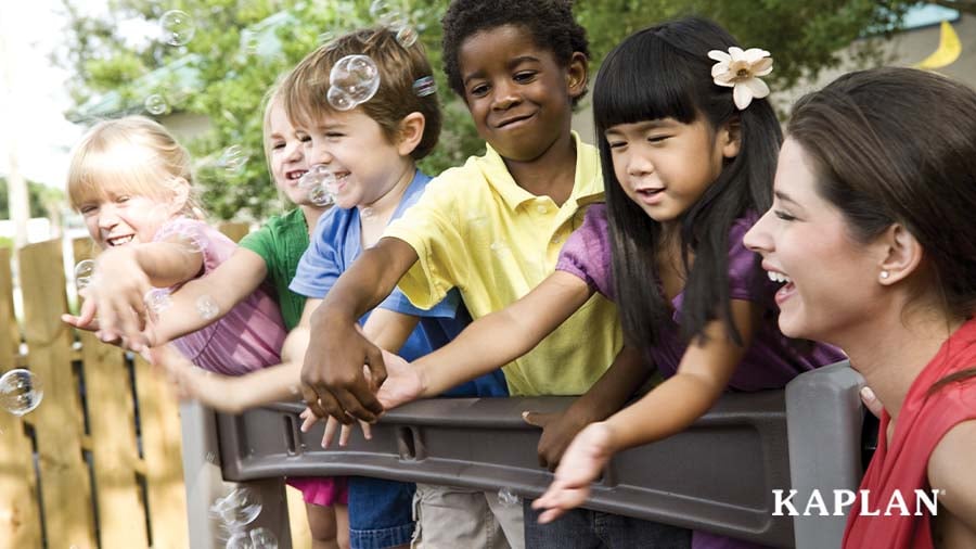 A group of young children and an adult stand outside, smiling as they try to catch bubbles which are floating in the air. 