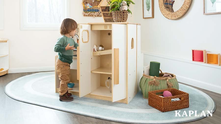 A young child stands near a Sense of Place for Wee Ones Sink and Refrigerator Kitchen set, a rounded Sense of Place rug covers the floor underneath. 
