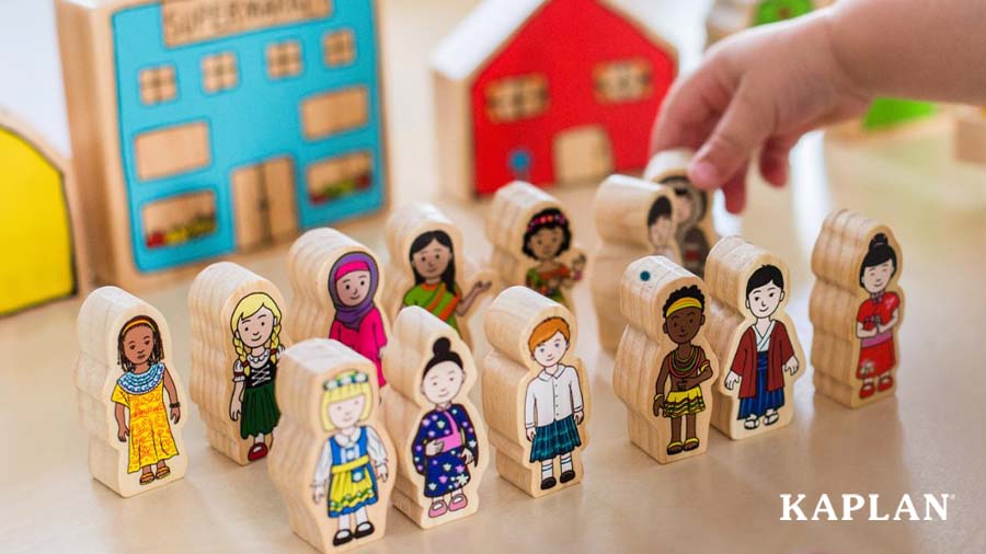 An image of Kaplan's Children From Around the World wooden blocks. A young child's hand is in the top corner of the image, touching one of the wooden block people. 