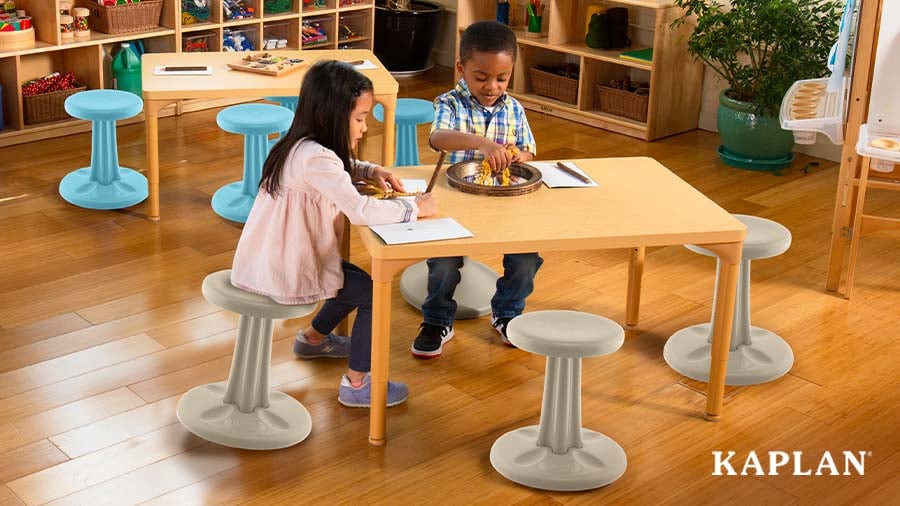 Two young children sit atop wiggle stools at a wooden classroom table as they work on writing and creative activities. 