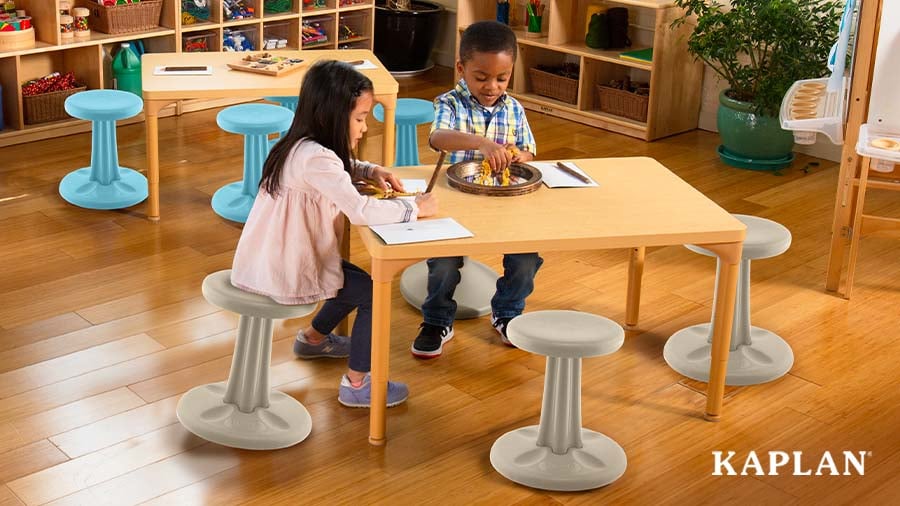 Two young children sit atop wiggle stools at a wooden classroom table as they work on writing and creative activities.