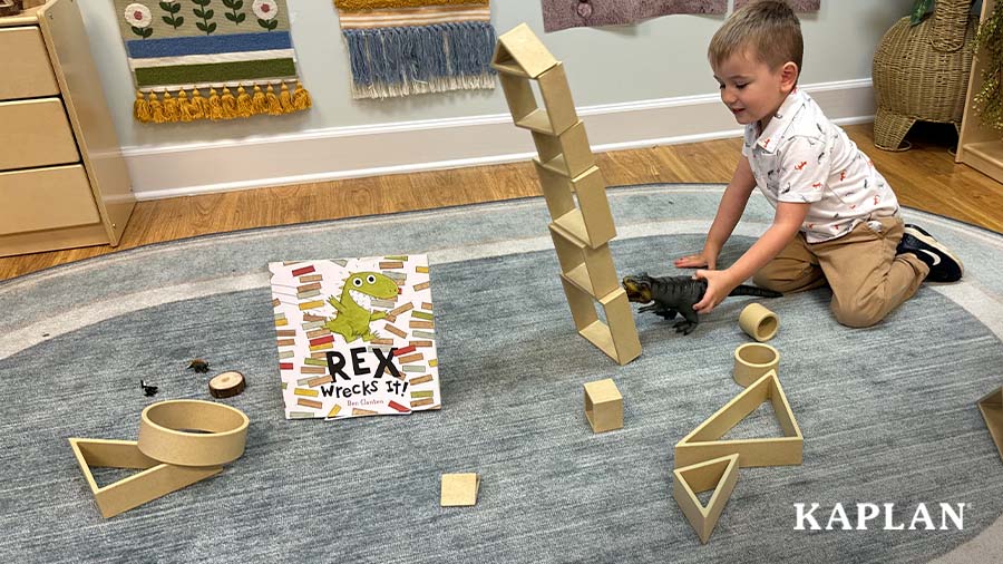 A young child is sitting on a blue classroom carpet, he is using a T-Rex toy to push over a tower of wooden blocks. 