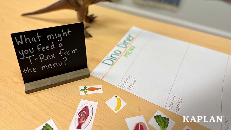 An image of the Dino Diner activity in action which features a hand-written menu and sign that says, "What might you feed a T-Rex from the menu?"