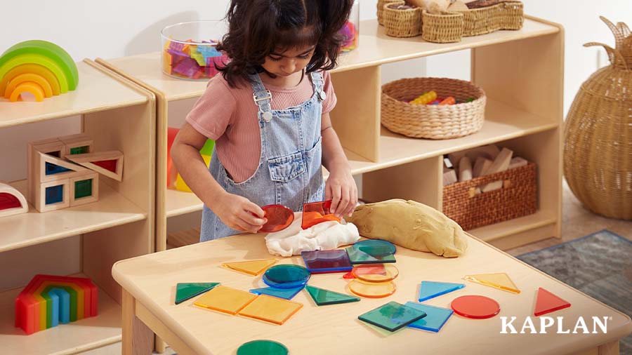 A young child stands beside a wooden table in a classroom block learning center. The child is holding two red plastic shape blocks while looking down at the table which contains a variety of colorful shape blocks. 