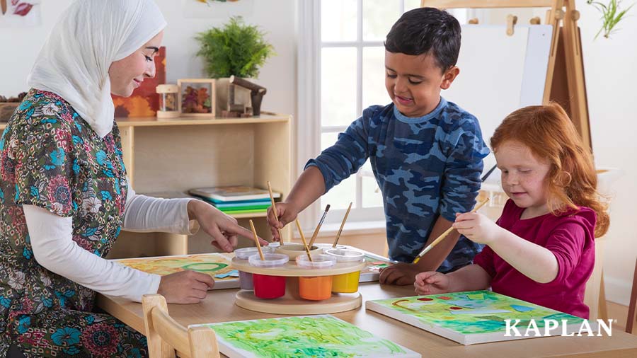 An early childhood teacher and two young children sit around a wooden table, they are painting on square canvas wall hangings. 