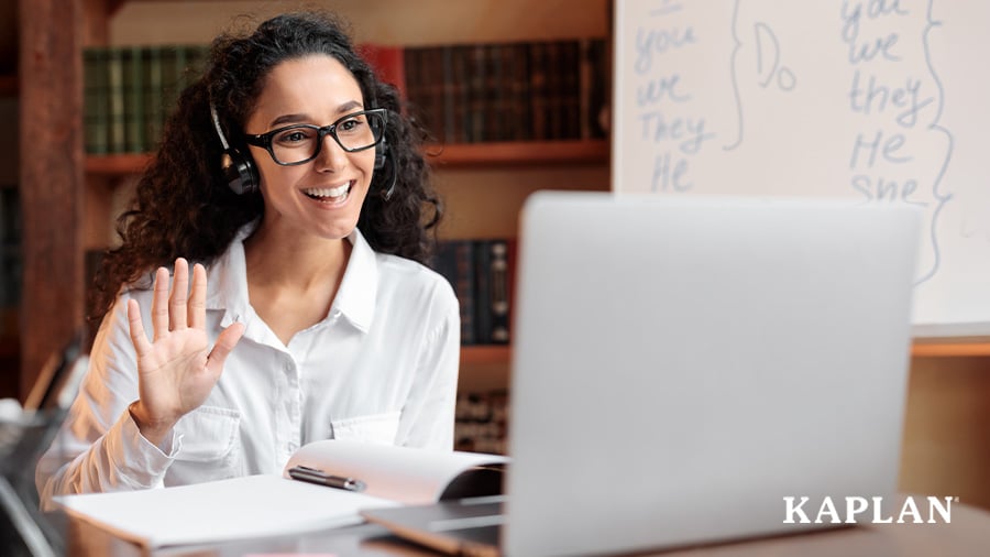 A woman smiles and waves her hand at an open computer which is on a desk in front of her. 