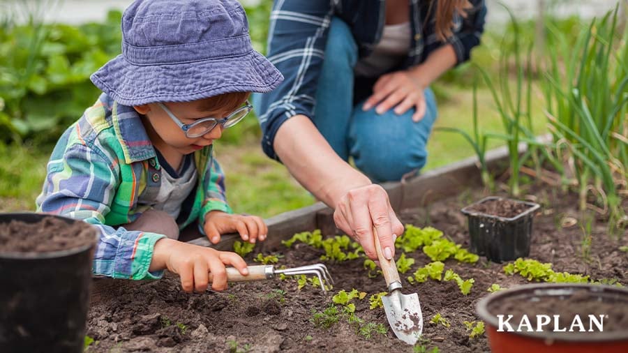A young child in a blue bucket hat uses a hand rake to move soil in a raised garden bed, an adult is pictured beside the child using a hand spade to also move the soil.