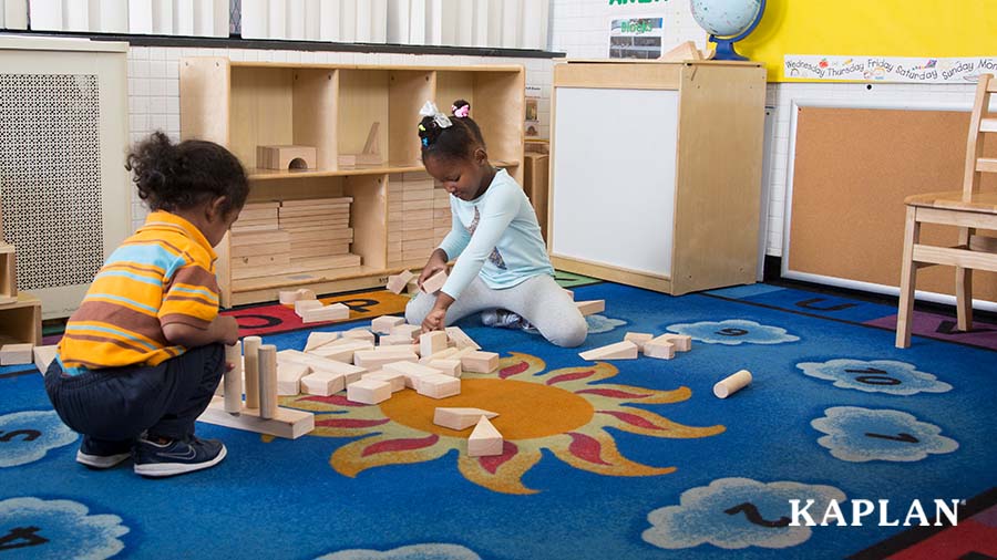 Two young children sit on the floor of an early childhood classroom, a pile of wooden blocks is on the floor between them. Each of the children is holding a block in one hand while placing it on top of a block which is laying on the floor.