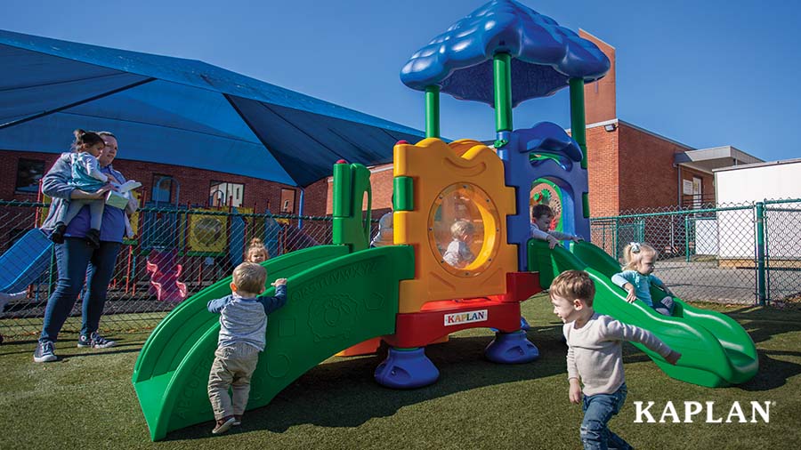 An early childhood teacher and young toddlers are outside on the playground. Several children are seen running, standing, and sliding down the play equipment. 