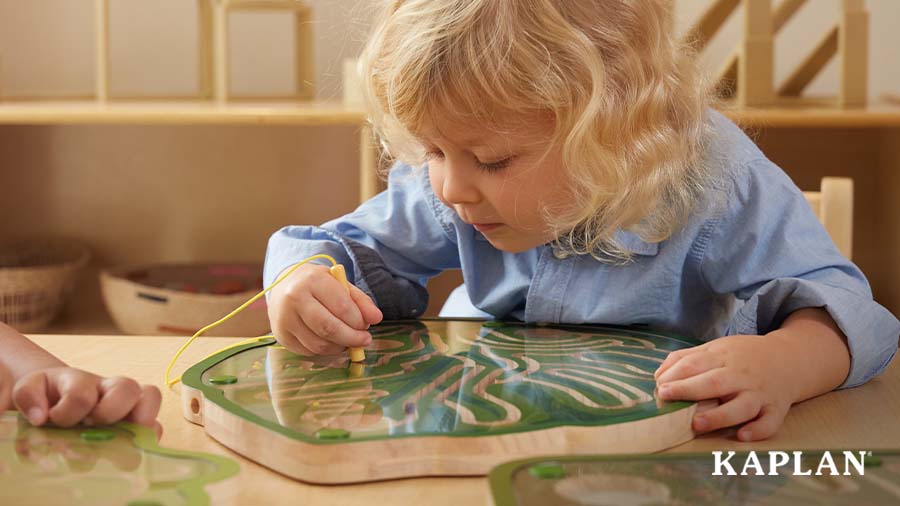 A young child in a blue shirt sits at a wooden classroom table, the child is using a magnetic wand to move a magnet marble through one of Kaplan's Leaf Maze puzzles. 