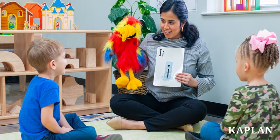 Early childhood educator sits on the floor with two young children, using a colorful bird puppet and a letter 'Ii' flashcard to lead a small group literacy lesson.
